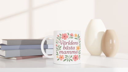 Mugg med texten Världens bästa mamma omgiven av mjuka blommor och blad i pastellfärger.